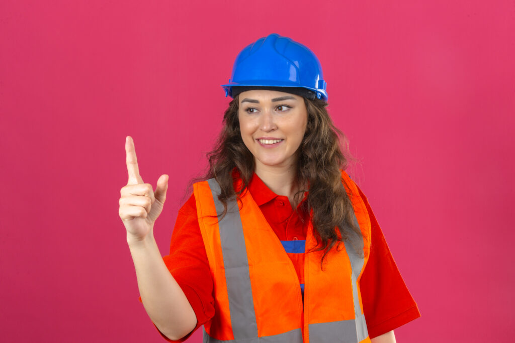 young builder woman in construction uniform and safety helmet smiling showing number one with fingers over isolated pink background